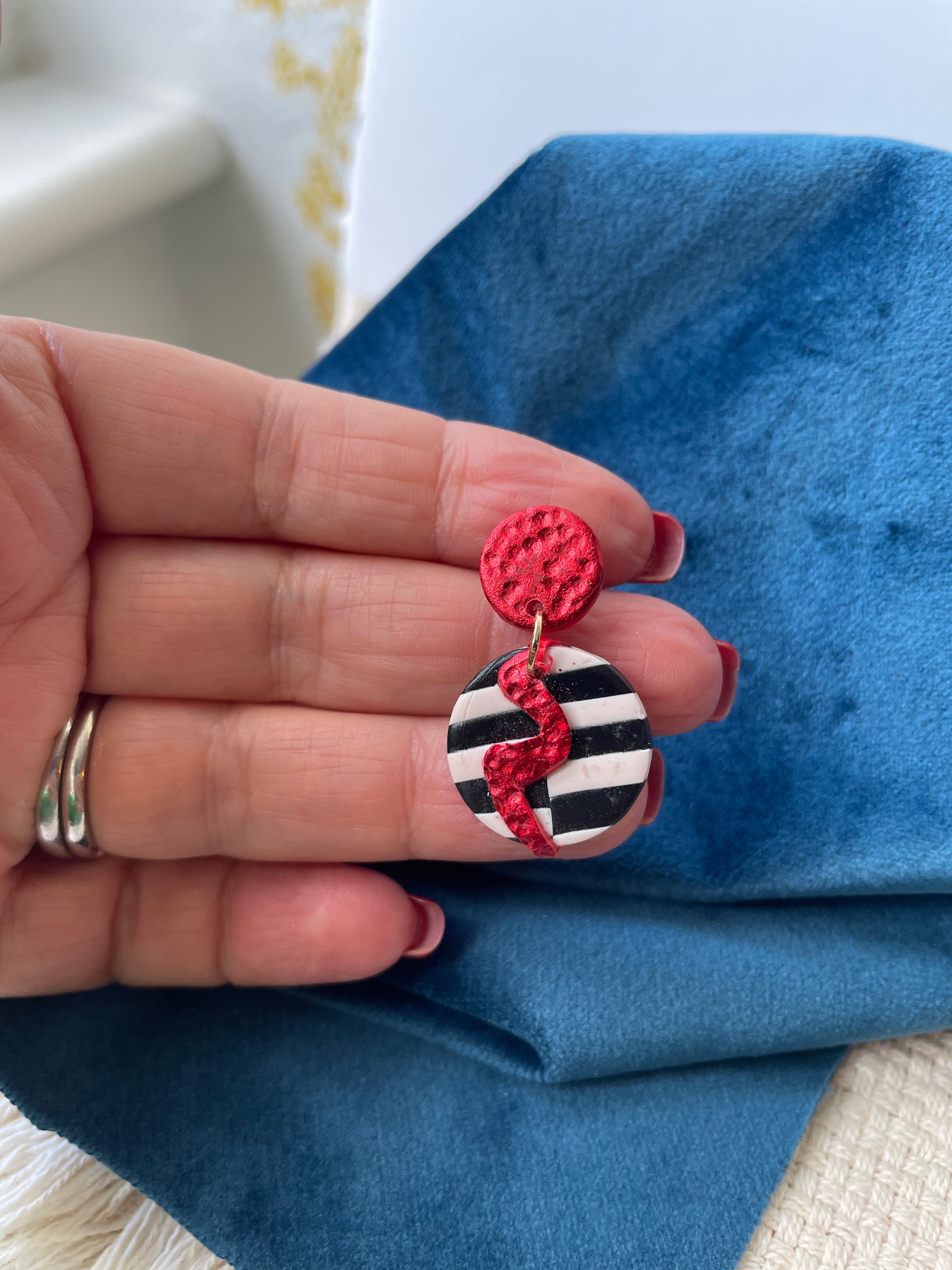 Spice up your look with these funky and vibrant earrings! Featuring a striking red lightning bolt on a bold black and white backdrop, these handmade beauties are sure to make a statement. Crafted from red clay and accented with shiny red foil, these earrings have a unique hammered texture.